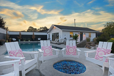 Backyard pool patio at sunset with white Adirondack chairs and pink patterned cushions around a blue-glass fire pit, facing a white single-story pool house