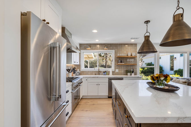 Sunlit modern farmhouse-style kitchen in a suburban home with a stainless-steel refrigerator and gas range, white cabinets, marble-topped island with brass pendant lights and sunflower centerpiece, hardwood floors and a garden-view window.