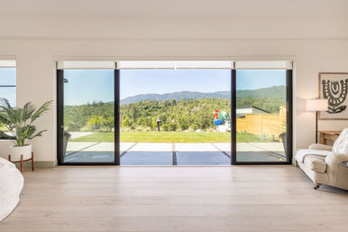 Bright modern living room with wide sliding glass doors opening to a backyard patio, green lawn, and panoramic tree‑covered hills beyond.