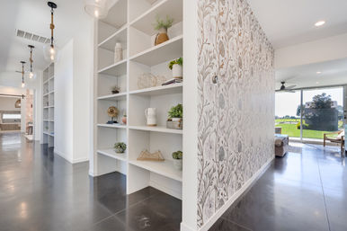 Modern open-concept hallway with white built-in shelving and decorative accents, floral patterned accent wall, polished concrete floors, pendant lights, and sliding glass doors with a sunlit backyard view.