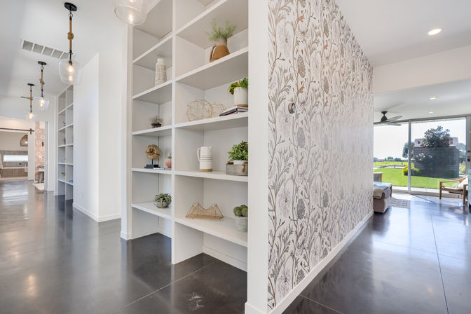 Modern open-concept hallway with white built-in shelving and decorative accents, floral patterned accent wall, polished concrete floors, pendant lights, and sliding glass doors with a sunlit backyard view.