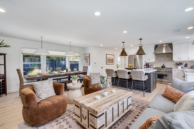 Sunlit open-concept living area and kitchen in modern farmhouse style with hardwood floors, gray sofa and orange pillows, two brown swivel chairs, rustic wooden trunk coffee table, long wooden dining table by large windows, island with woven barstools and pendant lights, and stainless steel range and refrigerator.