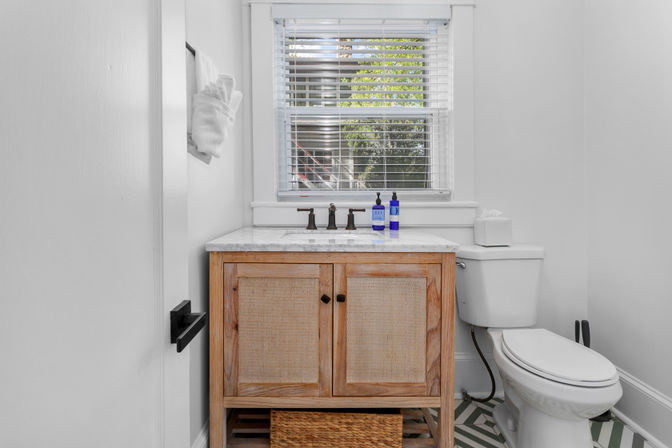 Sunlit small white bathroom with a natural wood vanity and marble countertop, oil-rubbed bronze faucet, blue soap bottles, window blinds, white toilet and geometric patterned floor tiles.