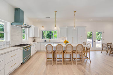 Bright modern farmhouse kitchen with white cabinetry, large island with five rattan-back bar stools, teal range and hood, brass hardware, glass globe pendants, light wood floors and French doors to backyard