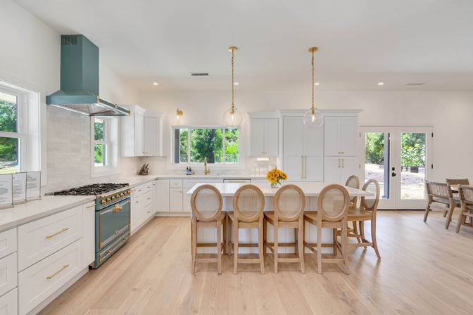 Bright modern farmhouse kitchen with white cabinetry, large island with five rattan-back bar stools, teal range and hood, brass hardware, glass globe pendants, light wood floors and French doors to backyard