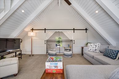 Cozy attic loft living area with white shiplap vaulted ceiling, sliding barn doors and black metal railing, gray sectional sofa with patterned pillows, pouf stools, floor lamp, TV, and board games stacked on a wooden coffee table.
