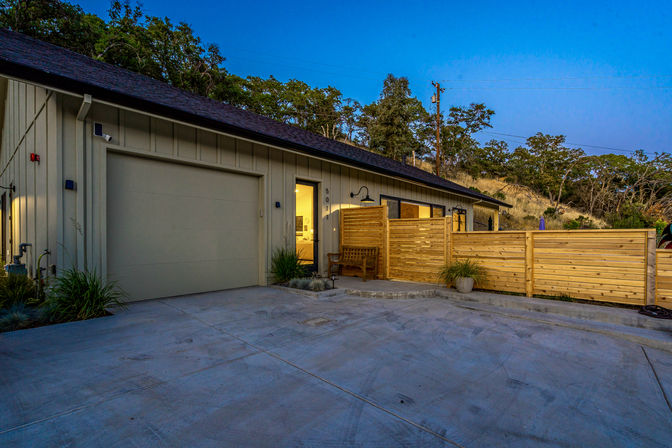 Dusk exterior of a modern single-story home with an attached garage, concrete driveway, and wooden privacy fence enclosing a small patio with a bench and potted plants, set against a tree-covered hillside.