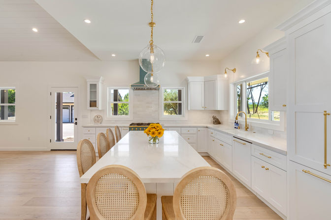 Bright open-concept modern kitchen with a long white marble island, rattan-back bar stools, brass fixtures and hardware, glass globe pendant lights, sunflower centerpiece and large windows overlooking a green yard.