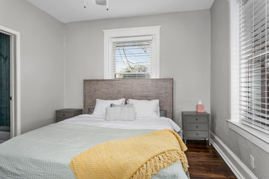 Cozy sunlit bedroom with rustic wooden headboard, neutral gray walls, white bedding and pillows, pale green quilt and cheerful yellow throw, matching gray nightstands, hardwood floors and window blinds.