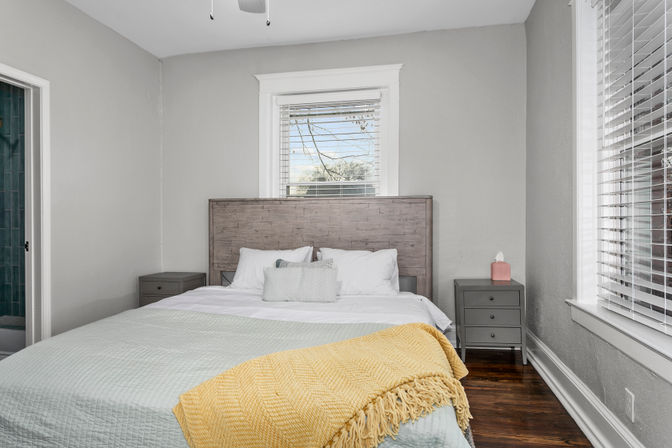 Cozy sunlit bedroom with rustic wooden headboard, neutral gray walls, white bedding and pillows, pale green quilt and cheerful yellow throw, matching gray nightstands, hardwood floors and window blinds.