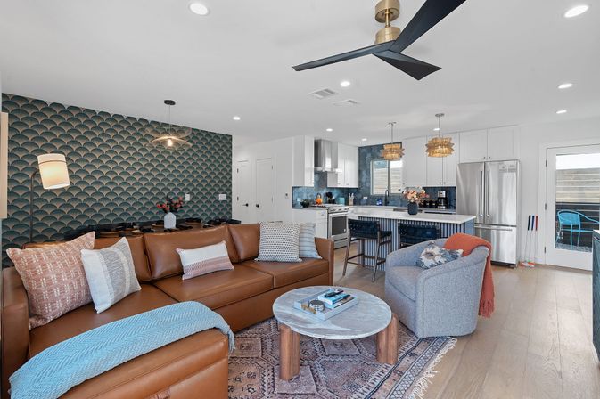 Stylish, cozy open-concept living room and kitchen with brown leather sectional, blue scallop-pattern accent wall, round marble coffee table, gray swivel chair, kitchen island with bar stools and wicker pendant lights, stainless-steel appliances, light hardwood floors and sliding patio door.