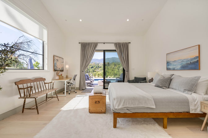 Sunlit modern master bedroom with king bed, wooden bench and desk, sliding glass doors opening to a pool and lush mountain view.