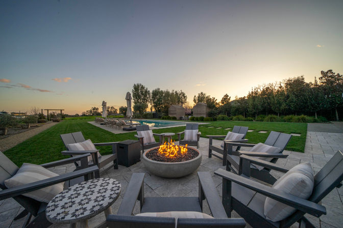 Sunset resort-style backyard patio with a glowing round fire pit surrounded by cushioned lounge chairs, a nearby pool with umbrellas, and a manicured lawn.