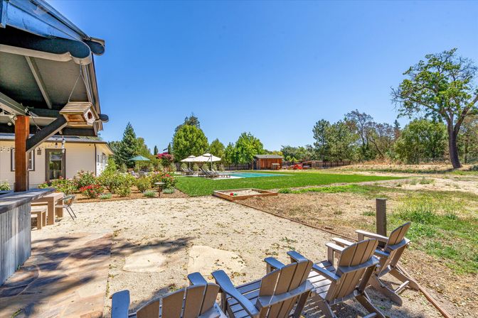 Sunny residential backyard with Adirondack chairs on a sandy patio, covered porch with a birdhouse, manicured lawn and pool area with lounge chairs and umbrellas, surrounded by trees under a clear blue sky.