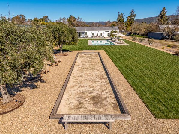 Aerial view of a sunny countryside backyard with a long bocce court and bench, gravel seating area under olive trees, manicured green lawn, rectangular swimming pool and a modern single-story house against blue sky and distant hills.