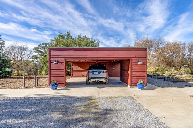 Sunlit red modern two-car carport with an SUV parked inside, flanked by blue ceramic planters with succulents, a concrete driveway and gravel forecourt, surrounded by trees under a bright blue sky with wispy clouds.