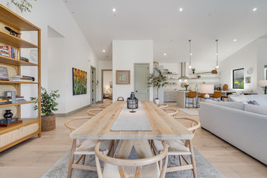 Bright, airy open-plan living room and kitchen featuring a long natural wood dining table with woven chairs, neutral sofa, kitchen island with tan bar stools and pendant lights, built-in shelving, plants and modern artwork.
