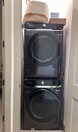 Stacked black front-loading washer and dryer in a narrow home laundry closet, space-saving setup with woven baskets and laundry detergent on top.