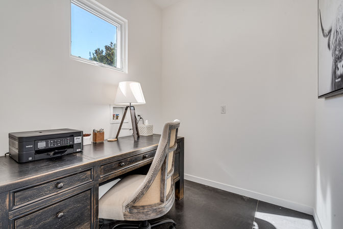 Minimalist home office with distressed dark-wood desk, beige upholstered swivel chair, compact printer, tripod table lamp under a small high window and bright white walls.
