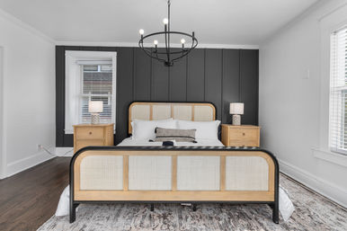 Contemporary bedroom with cane-front bed and black metal frame centered on a dark vertical paneled accent wall, wooden nightstands with lamps, round black chandelier, hardwood floors and patterned area rug under large windows.