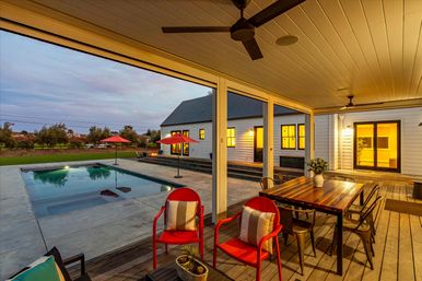Dusk-lit covered farmhouse patio with wooden dining table, two red chairs, ceiling fan, concrete swimming pool with red umbrellas and grassy yard beyond.