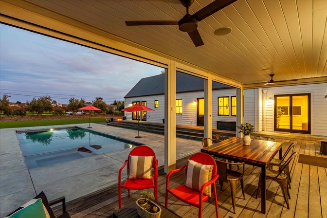 Dusk-lit covered farmhouse patio with wooden dining table, two red chairs, ceiling fan, concrete swimming pool with red umbrellas and grassy yard beyond.