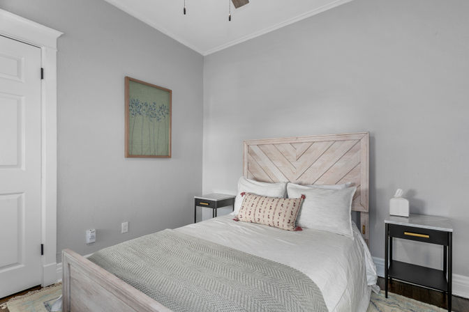 Cozy modern farmhouse bedroom interior with gray walls, light wood chevron headboard, white bedding, patterned lumbar pillow and black nightstands.
