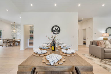 Bright open-concept modern farmhouse dining room set for eight on a long wooden table with woven placemats and floral napkins, light hardwood floors, minimalist white walls, adjacent cozy living area and patterned breakfast nook.