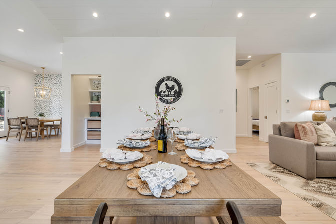 Bright open-concept modern farmhouse dining room set for eight on a long wooden table with woven placemats and floral napkins, light hardwood floors, minimalist white walls, adjacent cozy living area and patterned breakfast nook.
