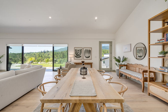 Bright, airy open-concept dining and living room with a long natural wood farmhouse table and round-back chairs, neutral sofa and bench seating, large sliding glass doors opening to lush green hills and distant mountain views, modern minimalist decor and abundant natural light.