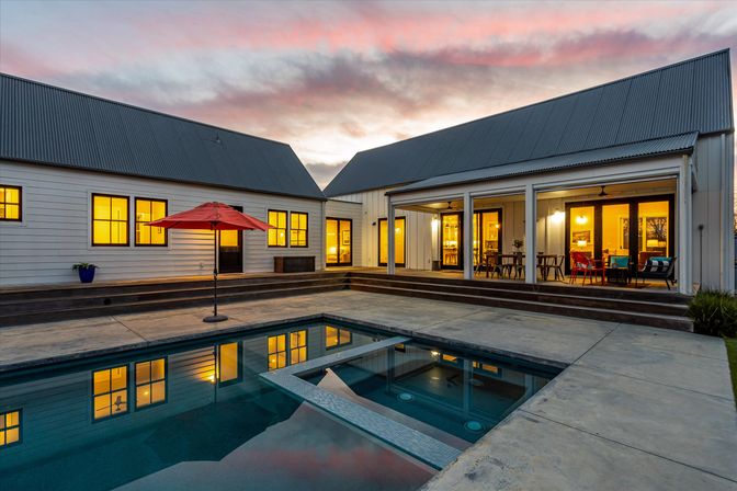 Modern farmhouse backyard at sunset with warm-lit windows, concrete patio, rectangular swimming pool with built-in spa, red patio umbrella, covered outdoor dining area and colorful seating reflected in the water.