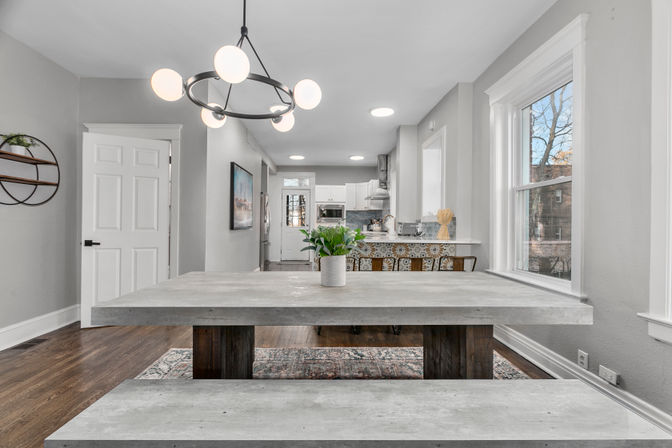 Open-plan dining area in an urban townhouse featuring a concrete-style farmhouse table and bench, globe chandelier, potted plant centerpiece, patterned-tile kitchen island, white cabinets, hardwood floors, and large windows.