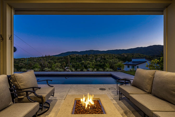 Dusk view from a cozy modern patio with cushioned outdoor seating, a lit rectangular fire pit, swimming pool, and rolling forested hills under a deep blue twilight sky.