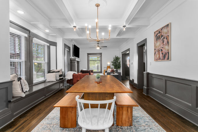 Sunlit open-concept dining room with a long wooden table and benches, white chair, built-in window seat under large windows, gray wainscoting, hardwood floors, brass chandelier, and adjacent living area.
