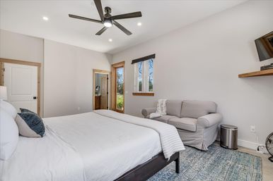 Bright modern master bedroom with white bedding, dark ceiling fan, gray loveseat, blue area rug, wood-trim window and wall-mounted TV.