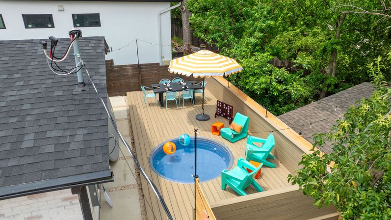 Rooftop wooden deck with a round plunge pool, yellow-striped umbrella, turquoise lounge chairs, outdoor dining table and string lights overlooking a leafy backyard