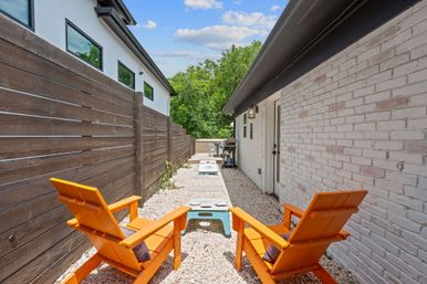 Narrow side-yard patio with two bright orange Adirondack chairs on gravel, cornhole boards along a stepping-stone path, modern white-brick house and horizontal wood privacy fence with a small outdoor dining area at the far end.
