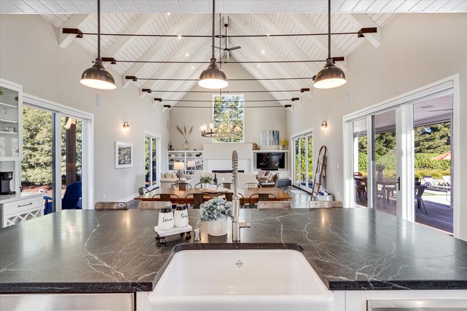 Bright open-concept kitchen and living room with vaulted white shiplap ceiling, industrial pendant lights, large dark marble island with farmhouse sink, long wooden dining table, fireplace and sliding glass doors opening to a sunlit patio and garden.