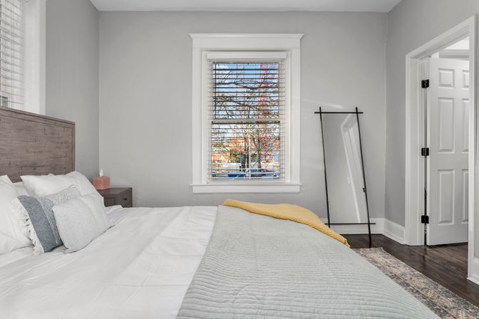 Sunlit modern bedroom with gray walls, wooden headboard and queen bed dressed in white and gray linens with a yellow throw, black-framed full-length mirror, hardwood floors and a window with blinds looking out to a tree-lined street.