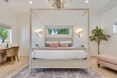 Sunlit modern bedroom with cream four-poster bed, crisp white bedding and pink accent pillows, wood floors, desk and chair by a window, potted green tree, blush upholstered bench, wall sconces and chandelier