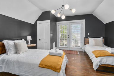 Cozy modern twin guest bedroom with white bedding and mustard-yellow throws, dark gray sloped walls, hardwood floors, pendant chandelier, bedside lamp and French doors with blinds letting in natural light.