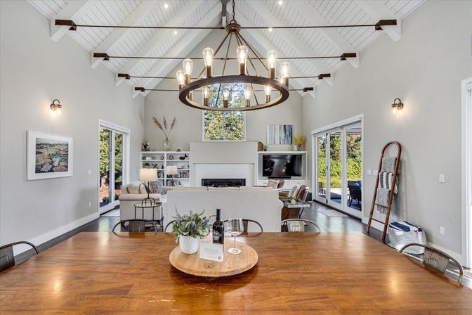 Open-plan modern farmhouse living and dining room with vaulted white shiplap ceiling and exposed beams, large circular iron chandelier, wooden dining table with wine and plant centerpiece in foreground, neutral sofas facing a fireplace and TV, built-in shelves and sliding glass doors opening to a sunlit backyard.