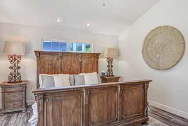 Bright master bedroom with large rustic wooden bedframe, neutral pillows, matching nightstands and ornate lamps, round woven wall decor and hardwood floor