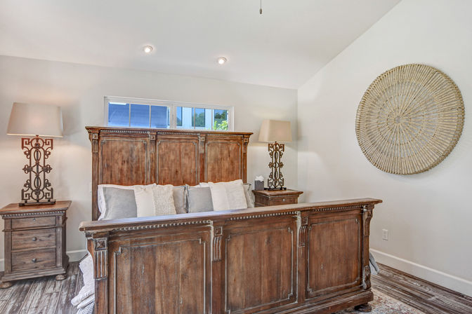 Bright master bedroom with large rustic wooden bedframe, neutral pillows, matching nightstands and ornate lamps, round woven wall decor and hardwood floor