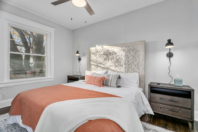 Cozy sunlit bedroom with ornate carved wood headboard, white bedding and coral throw blanket, dark wood nightstands with wall sconces, large window framing a tree, and ceiling fan overhead.
