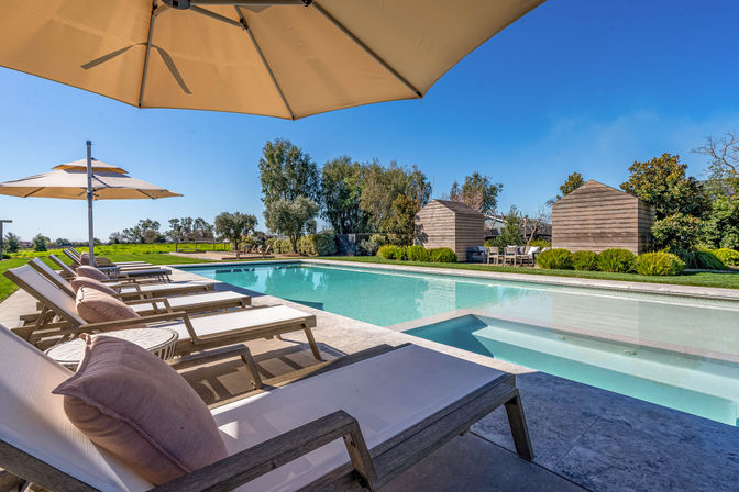 Inviting resort-style outdoor swimming pool with cushioned lounge chairs, oversized umbrellas, and wooden cabanas in a landscaped sunny garden.