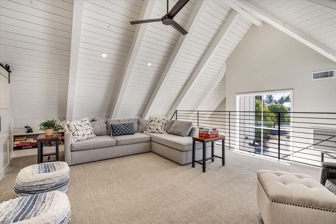 Cozy bright loft living room with vaulted white plank ceiling and fan, gray sectional sofa, side tables, poufs and metal railing overlooking treed yard