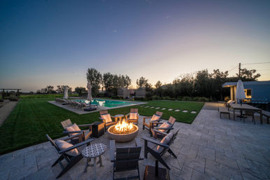 Twilight backyard oasis with a glowing circular fire pit surrounded by cushioned Adirondack chairs on a stone patio, next to a rectangular swimming pool, lounge chairs, manicured lawn and outdoor dining area.