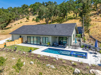 Aerial view of a sun-drenched hillside modern ranch home with a rectangular pool and spa, concrete patio with lounge seating and umbrellas, sliding glass doors, and oak-studded golden hills in the background.