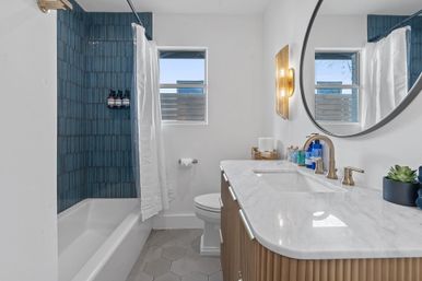 Bright modern home bathroom with blue vertical-tile shower and white tub, white shower curtain, hexagonal gray floor tiles, marble countertop on fluted wood vanity, round mirror, brass fixtures, small window and succulent decor.